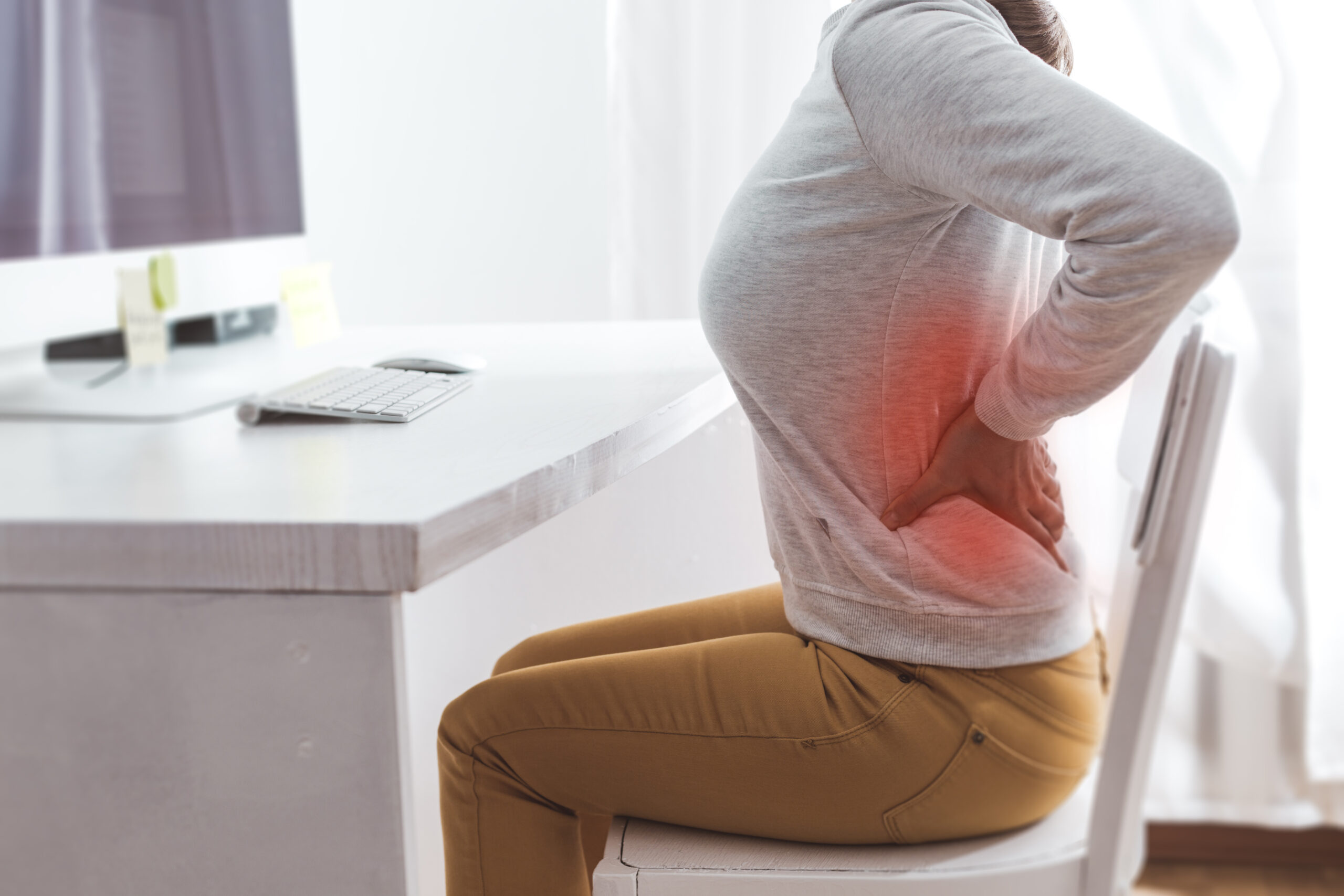 A woman sitting at her desk with a computer holding her back in pain