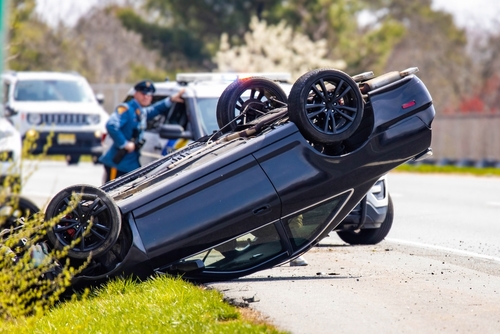 Overturned black SUV on the side of a highway as a police officer examines the accident scene in the background.