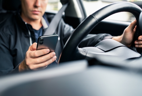 A man holds a smartphone in one hand and the steering wheel with the other, illustrating distracted driving.