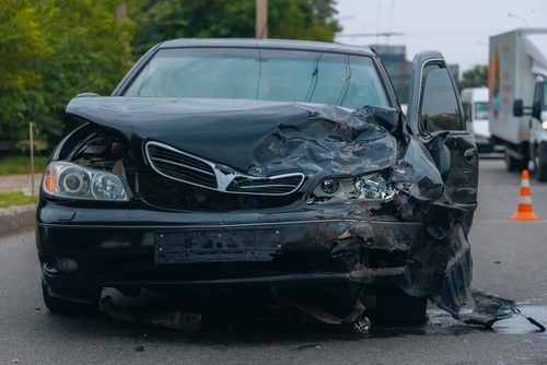 Black car with severe front-end damage after a collision, parked on the roadside with the driver’s door open.