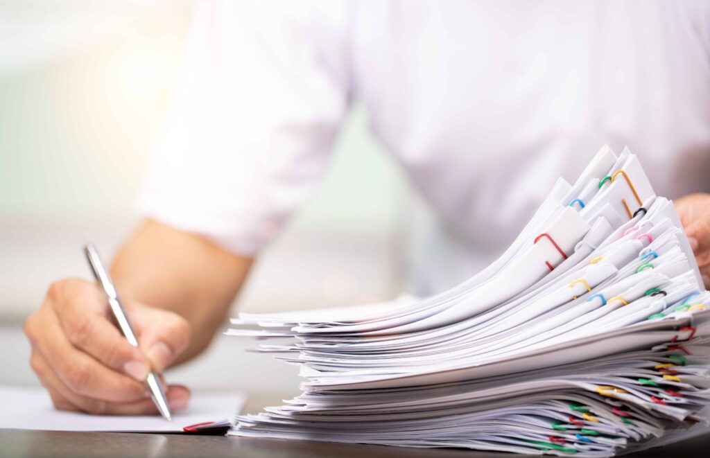 Person signing paperwork next to a large stack of legal documents clipped together, representing insurance claim or case file processing. Person signing paperwork next to a large stack of legal documents clipped together, representing insurance claim or case file processing.