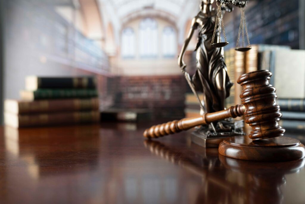Judge’s gavel on desk beside Lady Justice statue and law books in courthouse background.