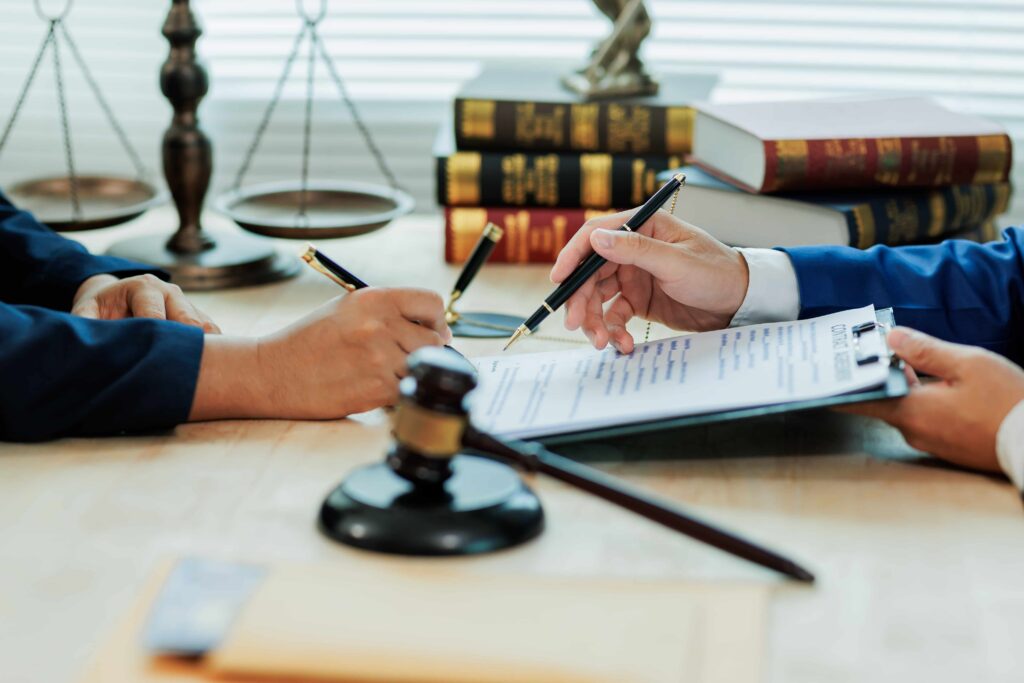 Lawyers reviewing and signing legal documents at a desk with a judge’s gavel, law books, and scales of justice in the background. Lawyers reviewing and signing legal documents at a desk with a judge’s gavel, law books, and scales of justice in the background.