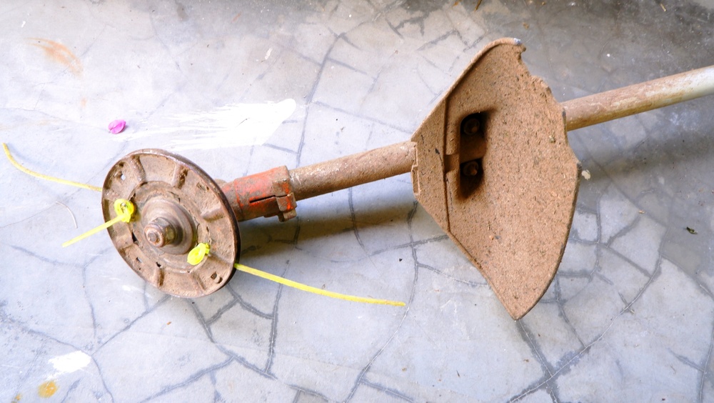 a close-up shot of the cutting head and damaged safety guard of a professional gasoline-powered grass trimmer. the orange plastic guard is visibly chipped and broken from wear and tear. the machine is resting on a cracked concrete floor, showing signs of heavy use with dust and dried grass debris. this image is suitable for topics relating to garden tool maintenance, equipment repair, workplace safety, and landscaping hardware.